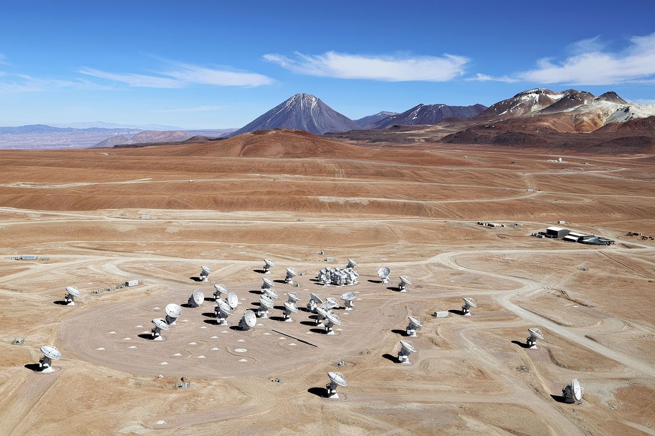 Aerial daylight photograph of white ALMA antennas spread across the dry Chajnantor Plateau, with mountains on the horizon.
