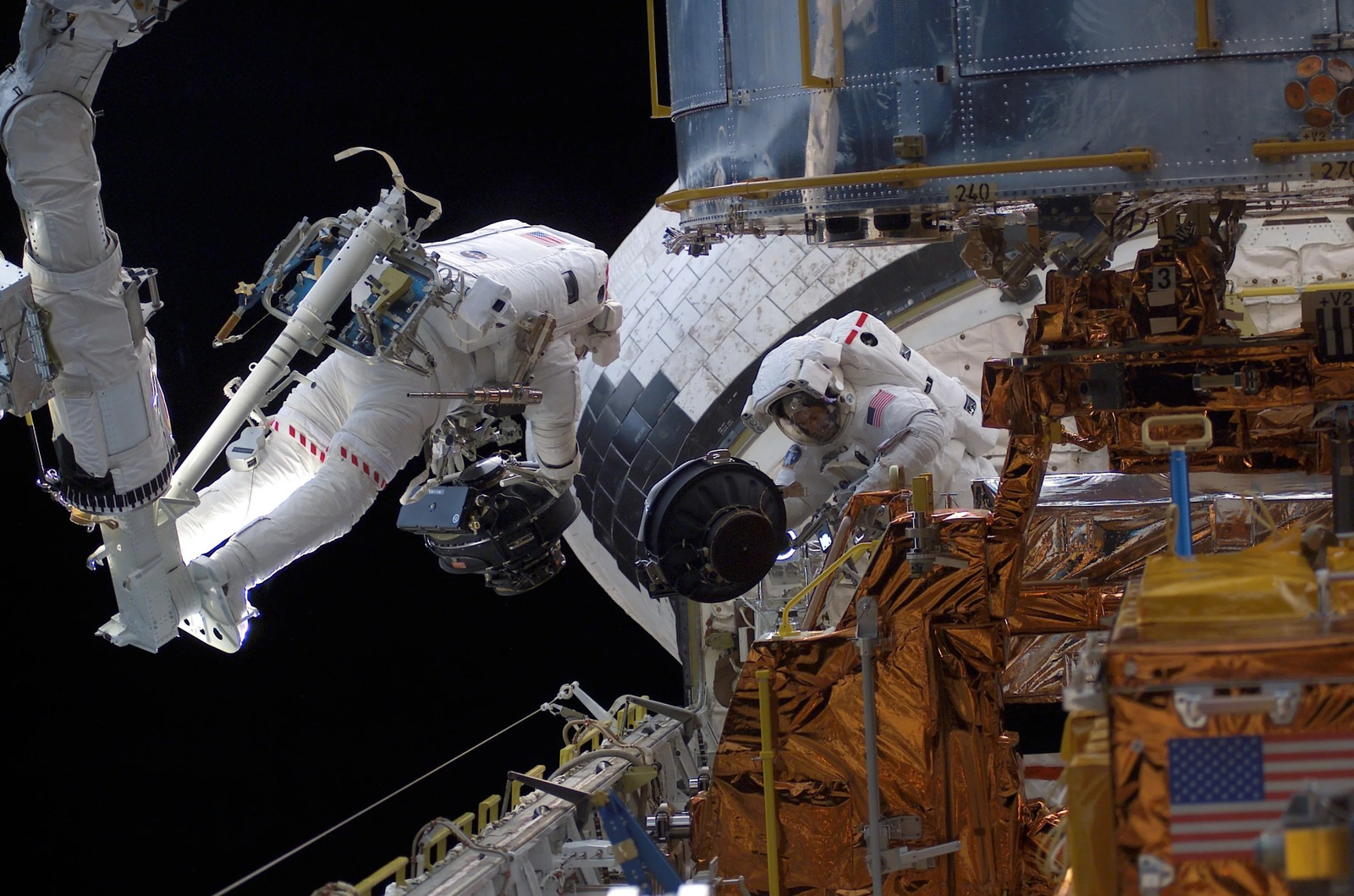 Two astronauts work beside Hubble, handling a large cylindrical reaction-wheel assembly amid the telescope's exposed equipment.